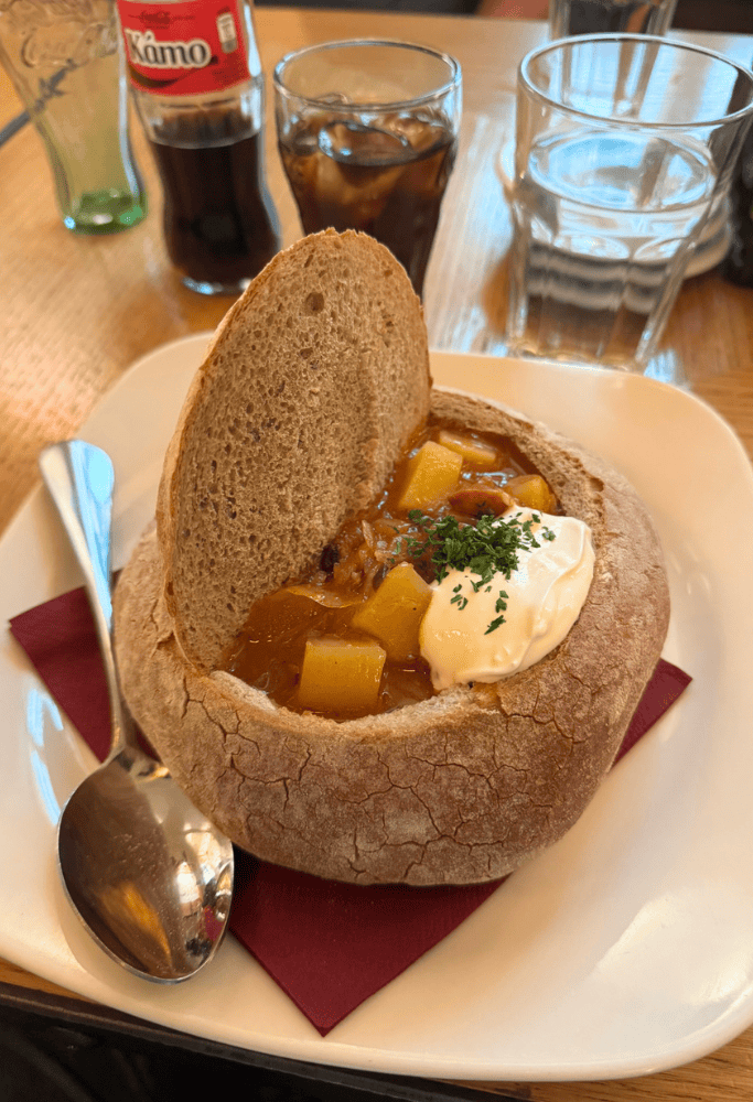 Traditional Czech goulash served inside a rustic bread bowl, topped with sour cream and herbs, alongside drinks at a Prague restaurant.