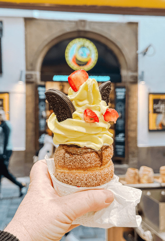 Chimney cake dessert topped with swirled cream, strawberries, chocolate spread, and chocolate cookies held inside a Prague food market during 3 Days in Prague.