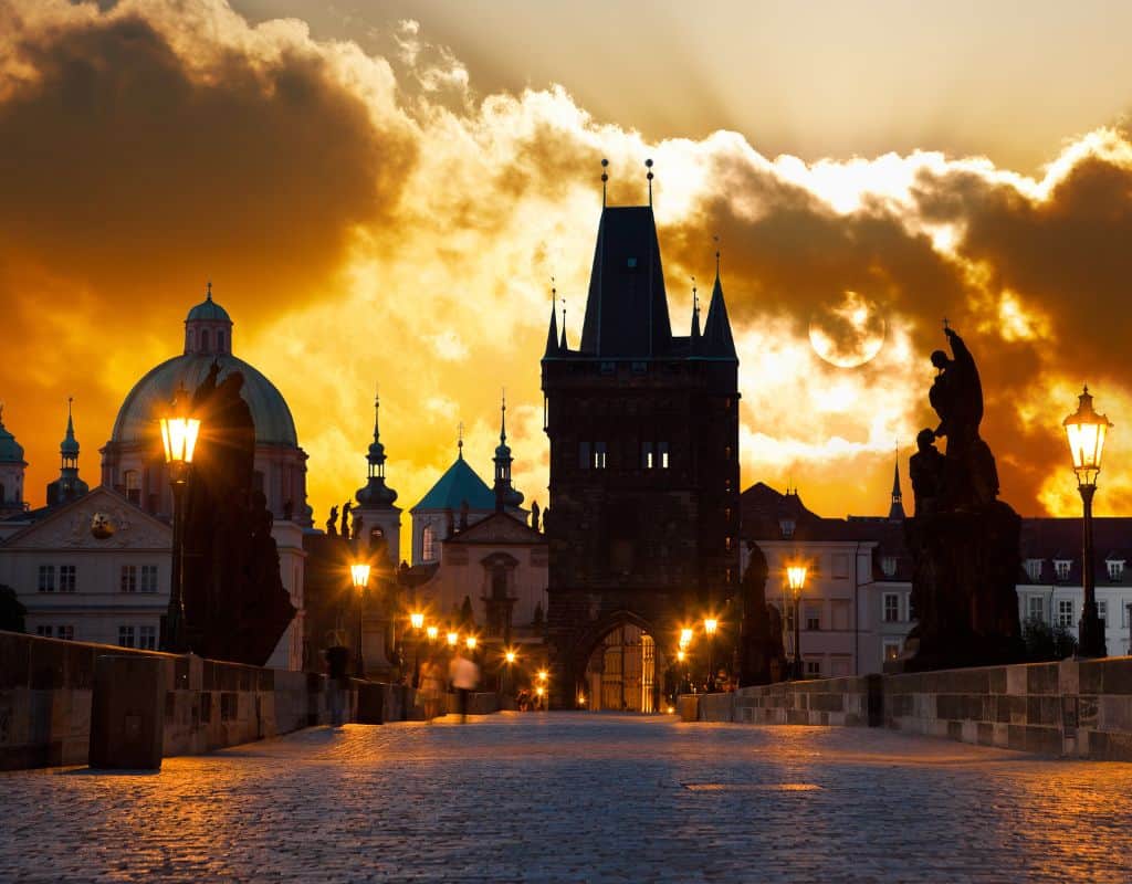 Charles Bridge at sunrise with glowing lanterns, dramatic golden clouds, and Prague’s historic towers silhouetted against the morning sky.